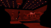 A dark, moody interior scene with oversized chess pieces arranged on a checkered floor, lit by many red ceiling lights. Silhouetted figures are visible in the foreground, and the overall color cast is strongly red.