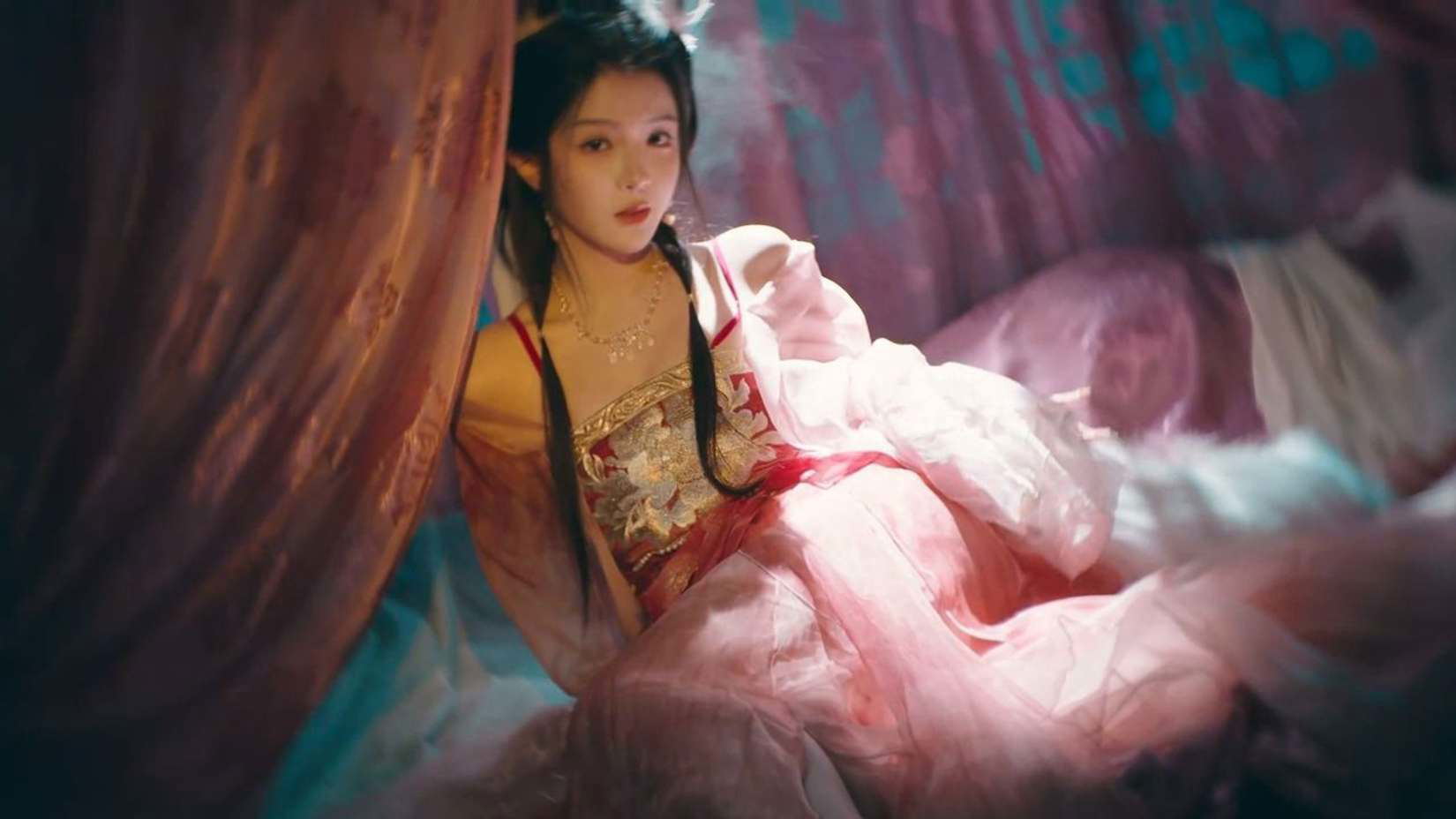 Person in traditional attire sitting in a softly lit room with pink and blue tones.