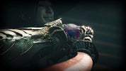 A close-up of an ornate metallic claw-like accessory with a glowing red and blue orb at the center, held by a person whose face is blurred in the dark background.