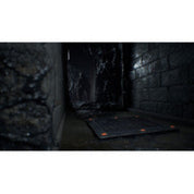 A dark, narrow stone corridor with a black rectangular floor mat or panel laid on the ground, shown in a dimly lit environment.