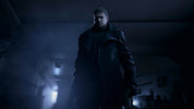 Man in a dark coat standing in a dimly lit room with a car in the background