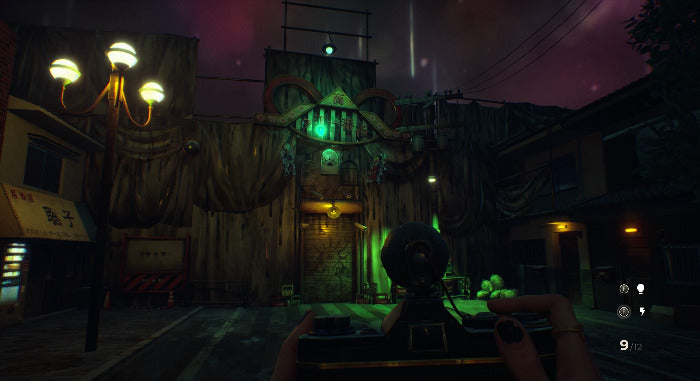 A dark, atmospheric video game scene showing a first-person view with a mounted camera or device in the foreground, facing a wooden structure with glowing green lights and a streetlamp on the left.