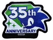 A 35th Anniversary embroidered patch featuring Sonic the Hedgehog artwork, with white '35th' text over green and blue sections, 'ANNIVERSARY' at the bottom, and a black stitched border. A small '©SEGA' mark is visible near the bottom edge.